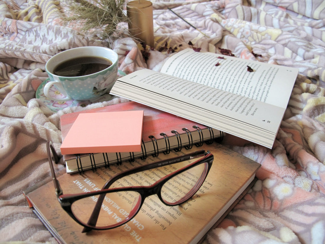 A book with a pair of glasses, journal, coffee, and notepad around it in a cozy space