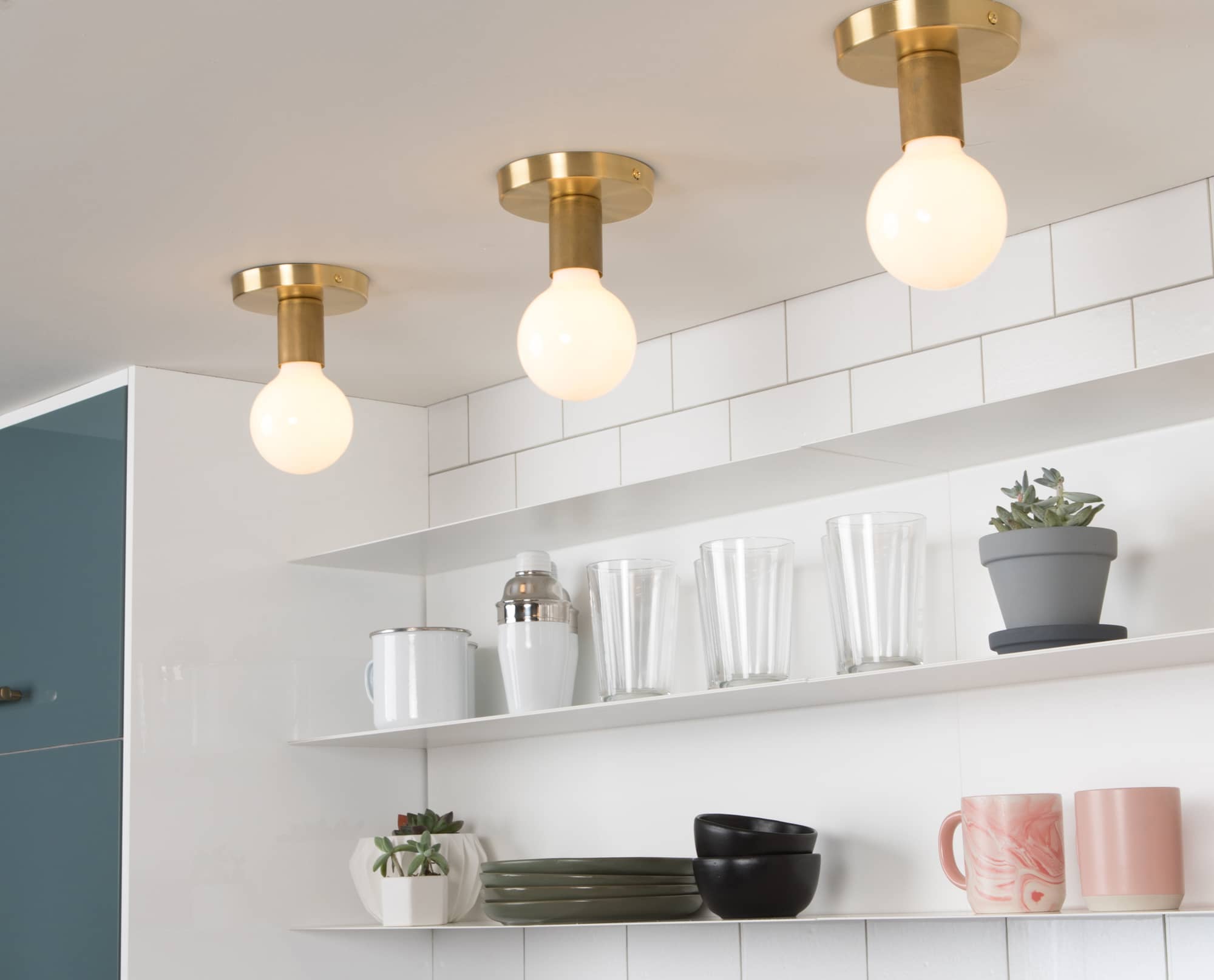 Button Lights in Raw Brass finish. Pictured with a G40 milk glass light bulbs. Mounted on ceiling over a kitchen counter and exposed shelving