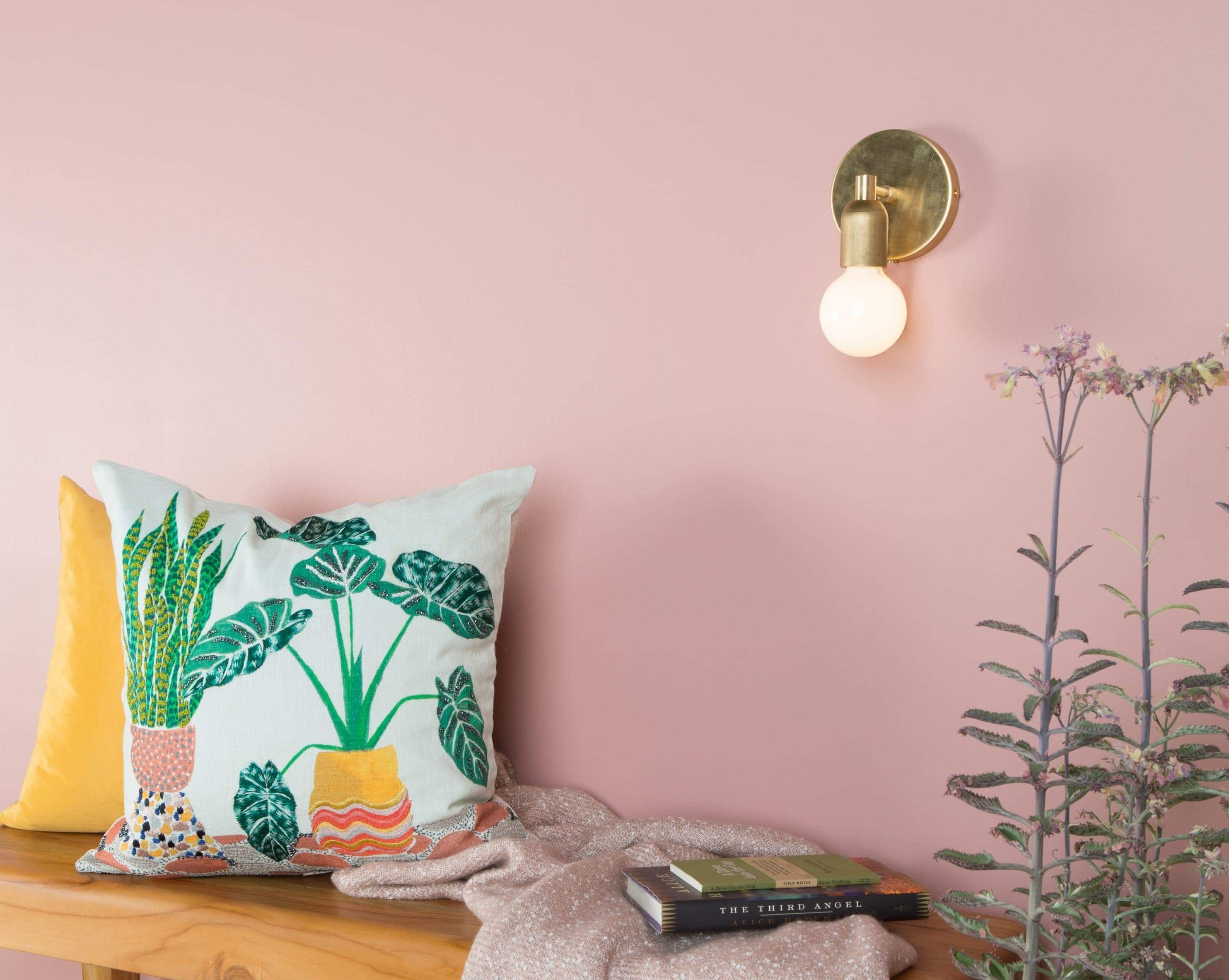 Room View of Junction Mini Solo Sconce in Raw Brass. Mounted on blush pink wall above teak bench with decorative pillows.